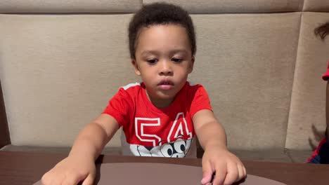 Cute-and-exotic-two-year-old-black-kid-waiting-impatient-for-his-food-in-a-restaurant,-wearing-red-shirt,-seated-next-to-mum