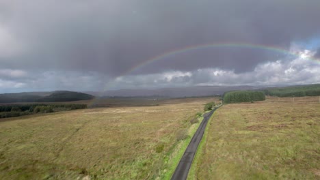 Aerial-drone-footage-in-the-rain-rising-up-above-a-long,-straight-road-looking-towards-a-birght-rainbow-with-dark-grey-clouds-in-the-background,-with-fields,-trees-and-moorland-in-Scotland