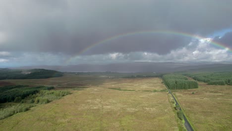High-aerial-drone-footage-in-the-rain-rolling-right-above-a-long,-straight-road-looking-towards-a-birght-rainbow-with-dark-grey-clouds-in-the-background,-with-fields,-trees-and-moorland-in-Scotland