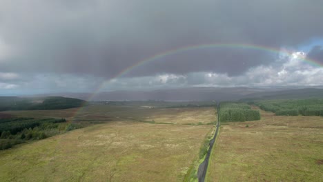 Aerial-drone-footage-descending-in-the-rain-above-a-long,-straight-road-looking-towards-a-birght-rainbow-with-dark-grey-clouds-in-the-background,-with-fields,-trees-and-moorland-in-Scotland