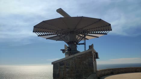 Radar-for-maritime-traffic-control-at-the-top-of-the-mountain-with-the-reflective-sea-in-the-background-on-a-sunny-summer-day,-shot-blocked-from-below