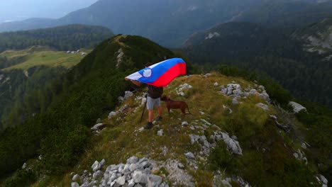 Forward-drone-footage-with-a-drone-passing-by-a-man-holding-a-Slovenia-flag-on-top-of-a-mountain-in-the-Alps-filmed-in-4k-in-nature-and-surrounding-mountains