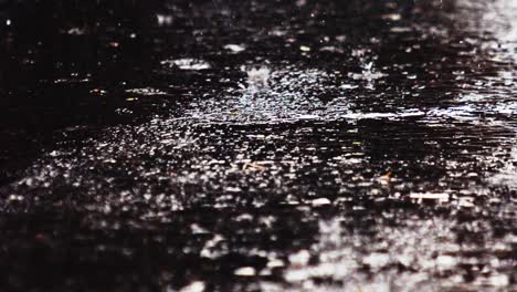Light-rain-water-droplets-falling-on-dark-asphalt-road,-close-up-ground-shot
