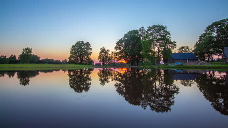 Timelapse-shot-of-sunset-in-the-background-and-cottage-beside-a-lake-in-the-foreground-at-dusk