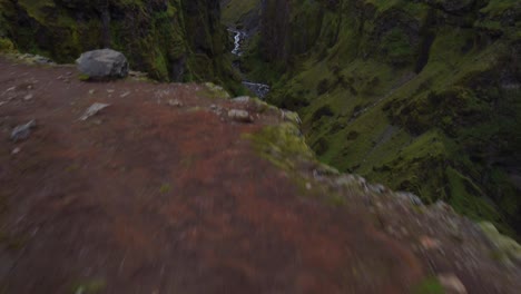 Canyon-waterfall-rising-quickly-drone-shot-of-red-jacket-hiker-admiring-view-of-green-canyon