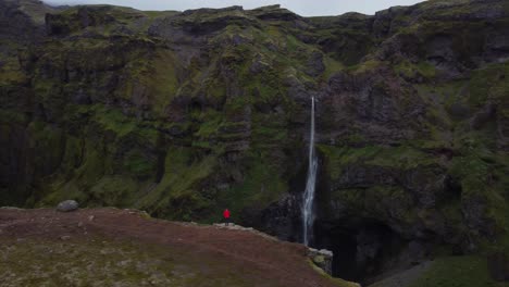 Canyon-waterfall-panning-drone-shot-of-red-jacket-hiker-admiring-view-of-green-canyon-and-cascading-waterfall