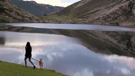 Young-woman-walking-the-dog-in-natural-stunning-scenery-mountains-lake-with-peak-reflection-in-still-water