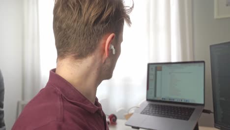 A-young-man-puts-wireless-earbuds-in-his-ears,-and-then-later-takes-them-out-while-he-is-working-at-his-home-office