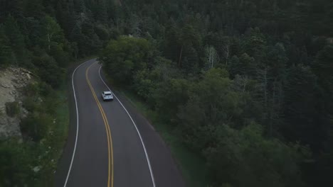 Drone-Shot,-Following-White-Car-Moving-on-Empty-Road-in-Evergreen-Forest-at-Dusk