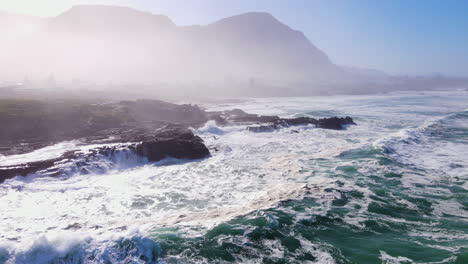 Wellen,-Die-Zerklüftete-Küstenlinie-Schlagen---Schäumendes-Meerwasser-Mit-Nebligem-Berghintergrund