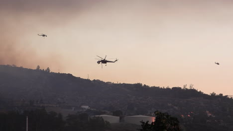 Helicopters-fly-through-smoke-by-wildfire-on-hills-in-California,-USA