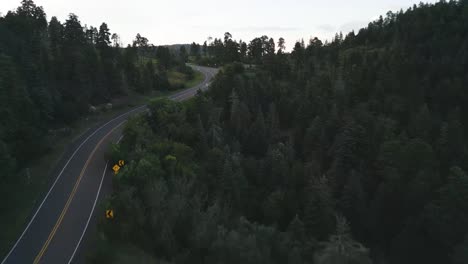 Drone-Aerial-View-of-White-Car-on-a-Road-in-Dense-Forest-Landscape-in-Twilight,-Sandia-Mountains,-New-Mexico-USA