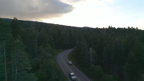 Drone-Shot-of-White-Car-Moving-on-Countryside-Road-Surrounded-by-Thick-Evergreen-Forest-in-Dusk