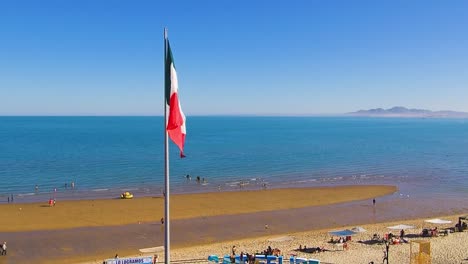 Blick-Auf-Eine-Drohne,-Die-Nach-Rechts-Fliegt-Und-Einen-Strand-Und-Die-Mexikanische-Flagge-Zeigt