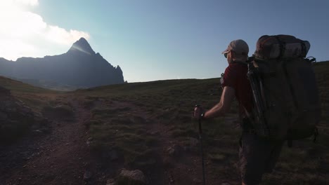 Back-view-of-hiker-with-big-backpack-walking-towards-mountain-peak-in-sunny-aftenoon-sunset-in-summer