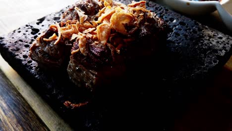 Closeup-Kobe-tenderloin-steak-self-cooking-on-a-square-lava-rock-steaming-hot-wooden-table-Japanese-traditional-sauce-bowl-background-rotating-360-degrees-room-for-text-below-4k60-topped-with-onions