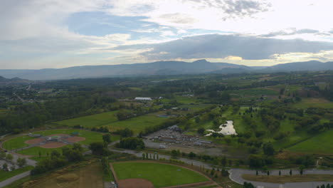 Aerial-view-of-fields,-wineries,-a-baseball-diamond,-lakes-and-mountains-outside-Kelowna,-on-a-summer-day