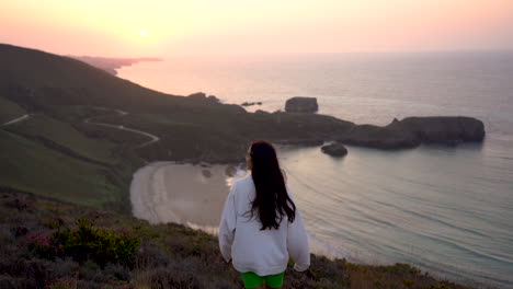 Young-woman-wearing-white-sweatshirt-hoodie-standing-on-top-of-windy-cliff-with-stunning-ocean-waves-view-at-sunset