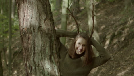 Young-woman-peeks-out-from-behind-a-tree,-puts-two-sticks-on-her-head-and-pretends-to-be-a-deer