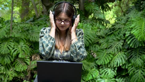 A-young-woman-adjusting-headphones-while-working-online-on-a-laptop-in-the-park-on-a-sunny-day