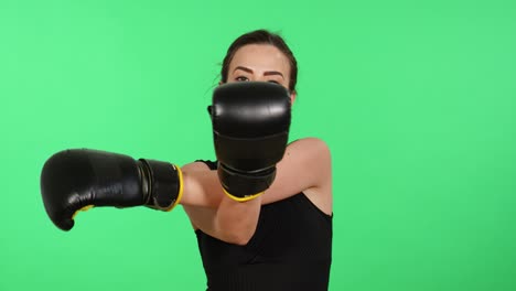 Attractive-brunette-woman-wearing-boxing-gloves-looks-toward-camera-while-stretching-arms-with-green-screen-in-background