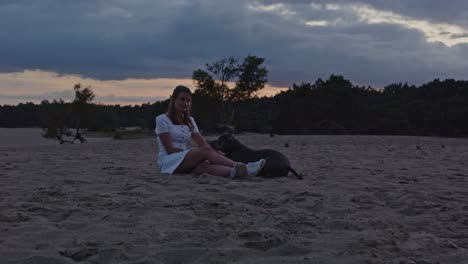 Wide-view-of-Young-woman-with-a-American-Staffordshire-Terrier-lying-next-to-her-in-sand-dunes-at-dusk
