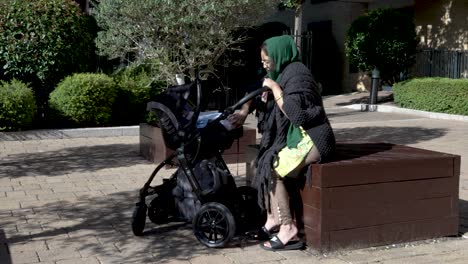 Eine-Junge-Asiatische-Muslimische-Mutter,-Die-Ihr-Neugeborenes-Baby-In-Einem-Kinderwagen-Tröstet,-Während-Sie-An-Einem-Sonnigen-Tag-In-Einem-Park-Im-Freien-Sitzt