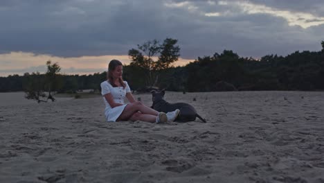 Wide-view-of-Young-woman-patting-a-American-Staffordshire-Terrier-while-sitting-in-sand-dunes