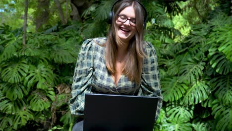 Happy-woman-with-headphones-laughing-while-using-a-laptop-in-the-green-park