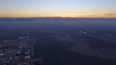 Aerial-view-of-fields-surrounding-small-town-with-sun-rising-above-the-mountains