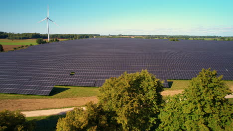 Aerial-view-flying-across-rural-farmland-above-treetops-overlooking-solar-panel-array-and-spinning-wind-turbine