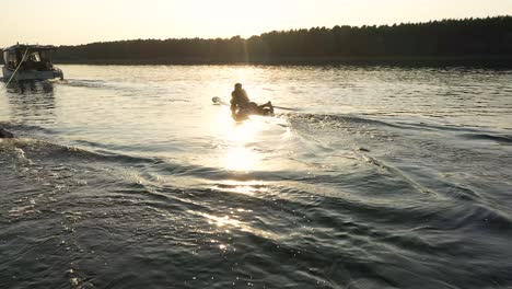 Surfer-Liegt-Auf-Surfbrett-Hinter-Boot-Mit-Sonnenuntergang