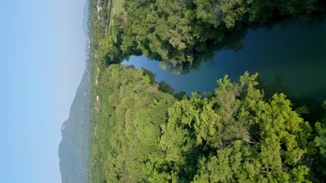 Aerial-vertical-flight-over-scenic-Rio-Munoz-River-during-sunny-day-and-blue-sky-surrounded-by-dense-forest-trees-at-shore---Mountain-landscape-in-backdrop
