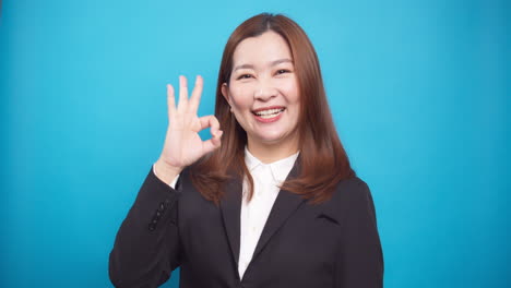 Beautiful-businesswoman-in-showing-ok-hand-gesture-smiling-standing-on-blue-background-looking-at-camera
