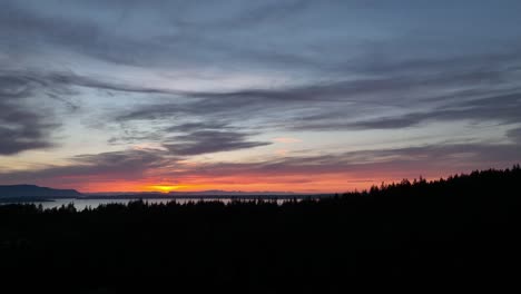 Rising-drone-shot-of-the-sun-setting-over-the-Pacific-Northwest-water-and-forest