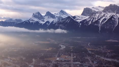 Slow-dramatic-left-pan-over-town-of-Canmore-in-a-valley-in-Alberta,-Canada,-as-the-sun-rises-over-low-clouds,-with-three-sisters-mountain-range-towering-in-the-background
