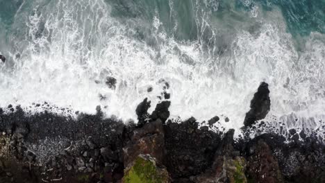 Foamy-waves-washing-pebbles-and-rocks-below-coastal-cliffs,-overhead