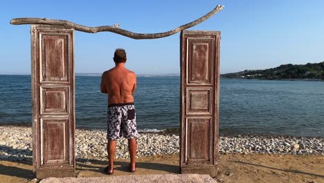 Decorative-seaside-wooden-door-and-back-view-of-man-standing-in-swimwear-leaning-against-doorframe