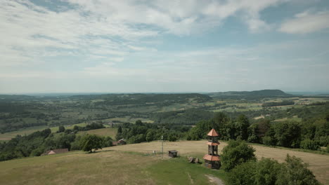 Drone-aerial-panoramic-landscape-shot-of-agricultural-fields-with-trees-and-forest-around-them-with-beautiful-blue-sky-and-hills-in-background-and-small-tower-in-the-top-of-the-hill-in-Slovenia