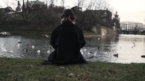 Young-woman-in-a-black-coat-sitting-in-a-park-in-autumn-in-afternoon-and-feeding-ducks-in-a-sea