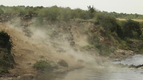 Clip-De-Cámara-Lenta-De-Una-Manada-De-ñus-Corriendo-Por-Una-Pendiente-Polvorienta-Y-En-El-Río-En-El-Masai-Mara,-Kenia