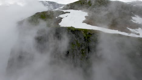 Aerial-view-of-dramatic-mist-revealing-cliff-edge-with-snow-patches-in-Simadal-Valley