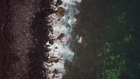 Top-down-aerial-relaxing-view-of-volcanic-rocky-beach-coast-while-waves-breaks-on-shore
