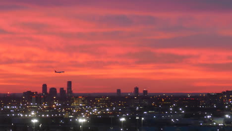 Silhouette-of-plane-landing-above-dusk-cityscape-with-glowing-lights-and-red-sunset-sky