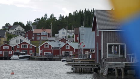 Statischer-Blick-Auf-Rote-Häuser-Am-Meer-Hinter-Einer-Schwedischen-Flagge,-Die-Unscharf-Weht