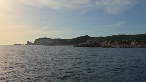Point-of-view-of-sailboat-bow-navigating-on-rippled-sea-water-toward-Corsica-island-cliffs-and-Capo-Pertusato-lighthouse-in-France,-50fps