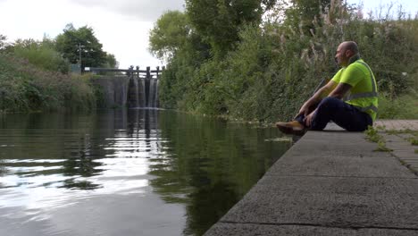 Warehouse-Worker-Taking-A-Break-To-Relax,-Sitting-At-The-Concrete-Edge-Of-The-River-Canal-In-Dublin,-Ireland