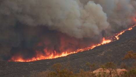 Wildfire-burning-along-a-mountain-slope,-with-black-and-grey-smoke-coming-up