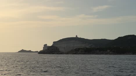 Point-of-view-of-sailboat-bow-navigating-over-rippled-sea-water-toward-Corsica-island-cliffs-and-Capo-Pertusato-lighthouse-in-France,-slow-motion