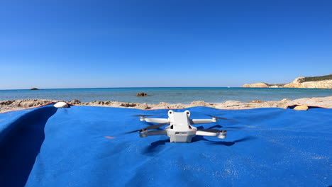 A-drone-begins-to-spin-its-propellers-on-a-towel,-on-a-Greek-beach-to-take-off